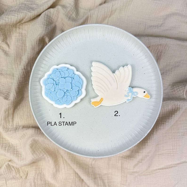 May include: Two PLA stamps on a light blue plate. One stamp is a blue hydrangea flower, and the other is a white goose with a blue bow. The text "1. PLA STAMP" is below the flower stamp.