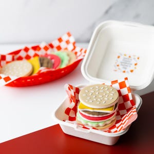 May include: A decorative hamburger cookie with sesame seed bun, cheese, lettuce, tomato, and a patty, presented in a white takeout container with red and white checkered paper. Additional cookies in a red basket.