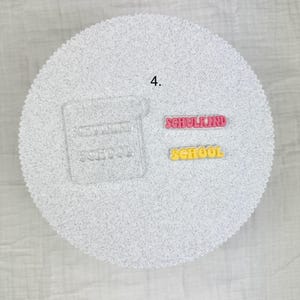 May include: A white, textured, circular surface with the number 4 in the upper left. Two colorful words are displayed: "SCHULKIND" in pink and "SCHOOL" in yellow. A clear, rectangular shape with the word "SCHOOL" is also visible.
