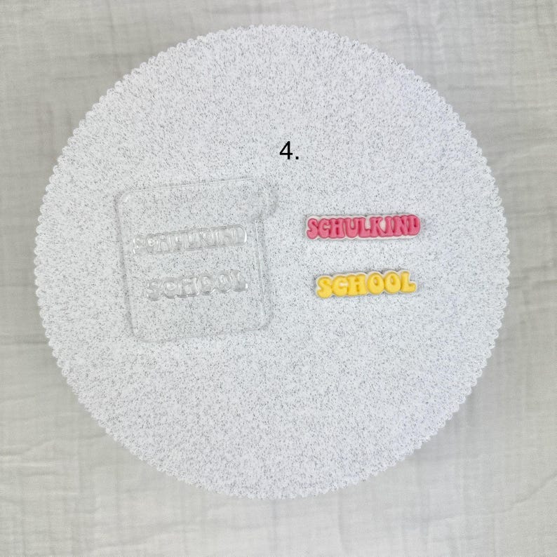 May include: A white, textured, circular surface with the number 4 in the upper left. Two colorful words are displayed: "SCHULKIND" in pink and "SCHOOL" in yellow. A clear, rectangular shape with the word "SCHOOL" is also visible.