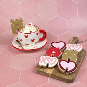 Puede incluir: Imagen temática de San Valentín con una taza blanca con corazones rojos, un asa roja y un platillo a juego. La taza está llena de crema batida y una galleta con forma de oso. Varias galletas con forma de corazón y labios están en una tabla de madera.