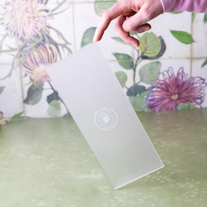 Pode incluir: Uma caixa retangular cinza claro com um logotipo circular que diz "Fabric Love With Handmade Cookie Cutter". A caixa é segurada por uma mão. O fundo apresenta um padrão floral com tons roxos e verdes.