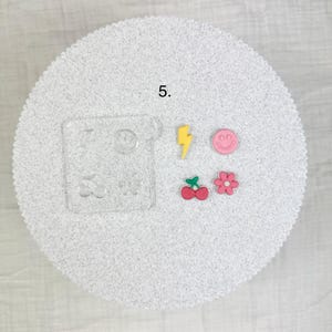 May include: A collection of colorful resin charms and a clear acrylic mold. The charms include a yellow lightning bolt, a pink smiley face, a red cherry, and a pink flower. The mold has various shapes, including a lightning bolt, a smiley face, a heart, and a flower.