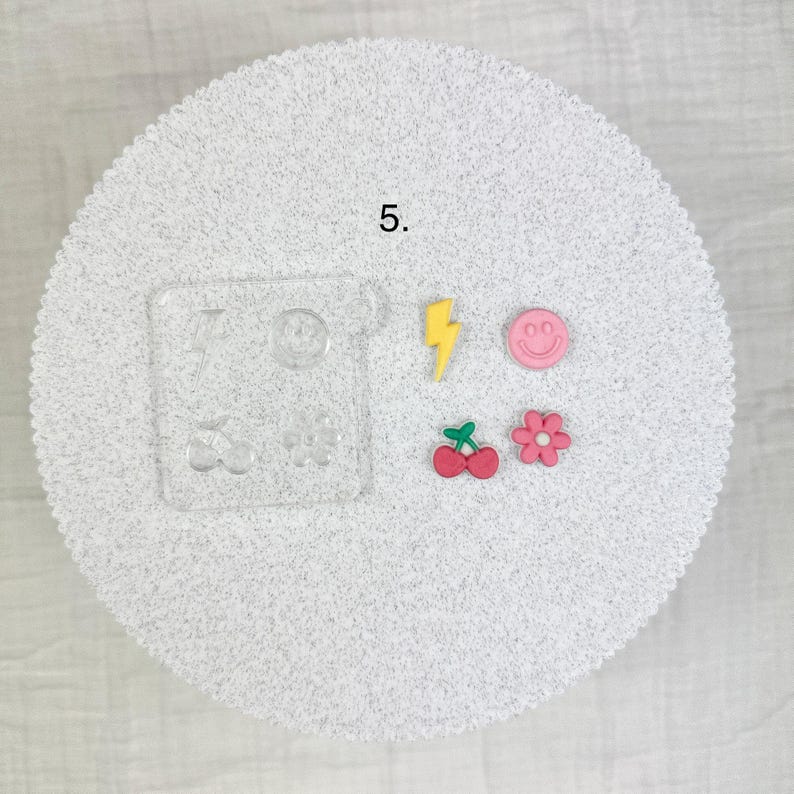 May include: A collection of colorful resin charms and a clear acrylic mold. The charms include a yellow lightning bolt, a pink smiley face, a red cherry, and a pink flower. The mold has various shapes, including a lightning bolt, a smiley face, a heart, and a flower.