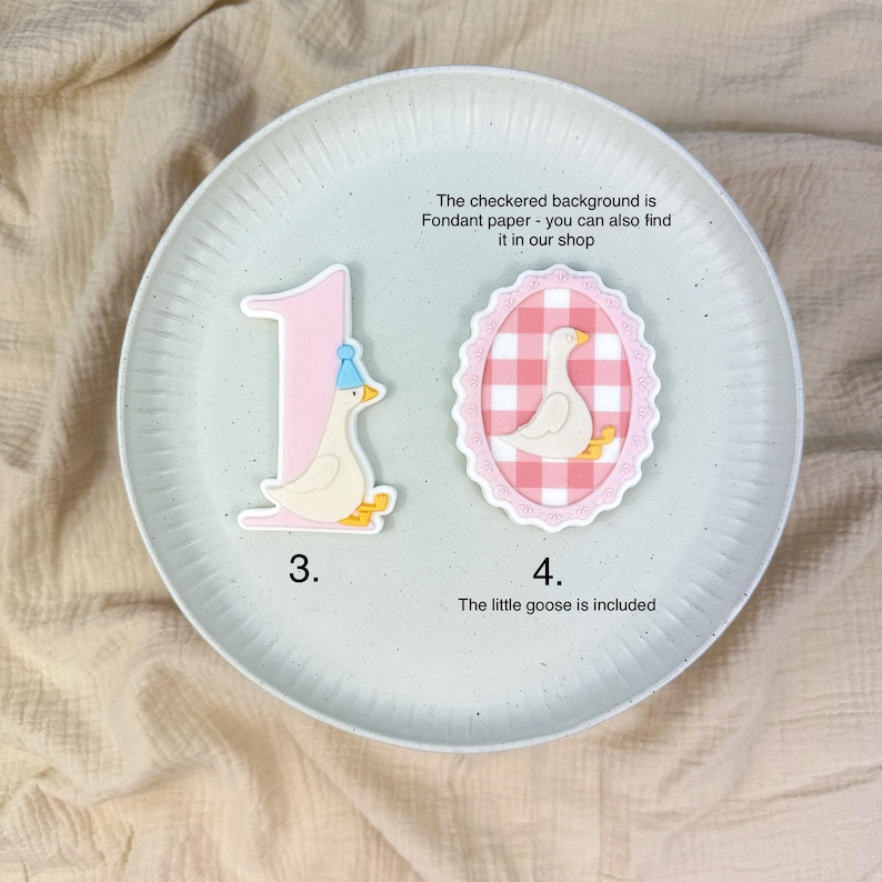 May include: A light pink number one and a white goose with a blue party hat are on a white plate. A second goose image is inside a pink oval with a checkered background. The text says, "The little goose is included."
