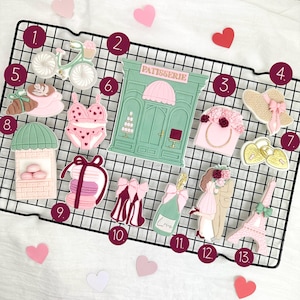 以下が含まれることがあります： バレンタインデーをテーマにしたクッキーのコレクション。自転車、"PATISSERIE"の文字が入ったお店、花の付いたハンドバッグ、麦わら帽子、クロワッサン、ビキニ、マカロンタワー、シャンパンボトル、カップル、エッフェル塔など。