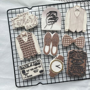 May include: A collection of nine decorated sugar cookies on a black wire rack. The cookies feature various designs including a bow tie, a vest, a watch, a credit card, a glass of whiskey, and a message that says "Happy Father's Day".
