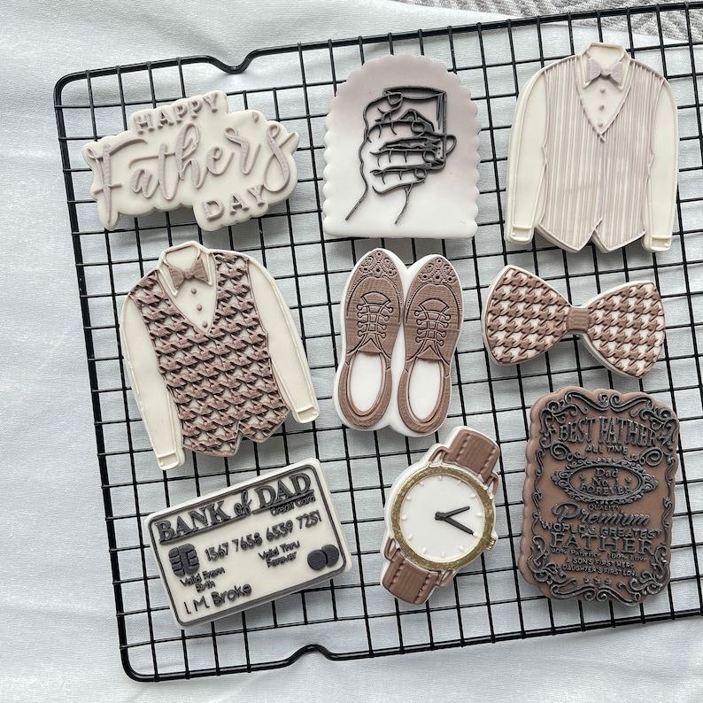 May include: A collection of nine decorated sugar cookies on a black wire rack. The cookies feature various designs including a bow tie, a vest, a watch, a credit card, a glass of whiskey, and a message that says "Happy Father's Day".