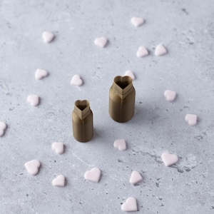Op de afbeelding: Twee bruine, hartvormige koekjessnijders met een hartvormige opening aan de bovenkant. Kleine, lichtroze hartvormige snoepjes zijn verspreid rond de uitstekers op een grijze ondergrond. De uitstekers zijn gemaakt van een stijf materiaal.