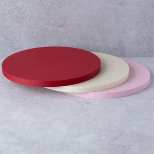 Pode incluir: Três tábuas redondas de madeira nas cores vermelho, creme e rosa estão empilhadas. As tábuas têm um acabamento liso e fosco e estão dispostas de forma ligeiramente deslocada, mostrando suas diferentes cores e texturas. As tábuas são provavelmente usadas para servir ou exibir.