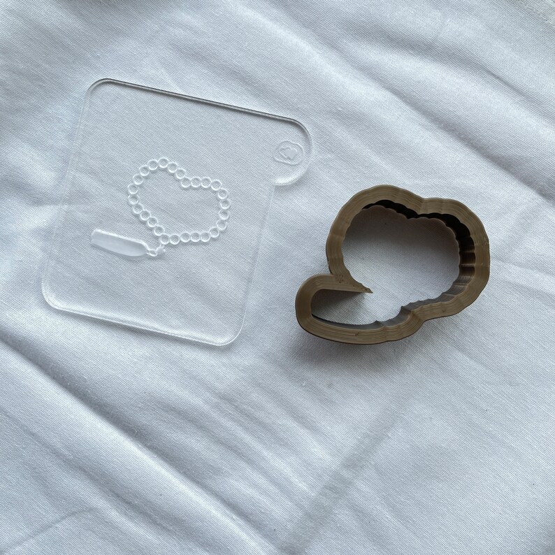 May include: A clear plastic template with a design of a necklace and a small rectangle. Next to it is a brown plastic cookie cutter shaped like a cloud.