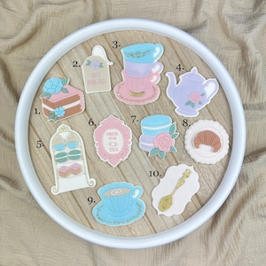 Könnte beinhalten: Eine Sammlung skurriler Dekorationen zum Thema Teeparty in Pastellfarben. Enthält einen Kuchen, gestapelte Teetassen, eine Teekanne, Makronen, ein Croissant und eine Teetasse. Auf einem weißen Tablett angeordnet.