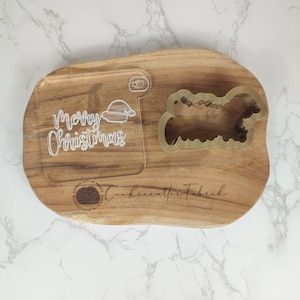 May include: A wooden cutting board with a "Merry Christmas" inscription and a Santa hat design. The board also features a cookie cutter in the shape of a Christmas tree. The text "Cookiecutter Fabrik" is engraved on the board.
