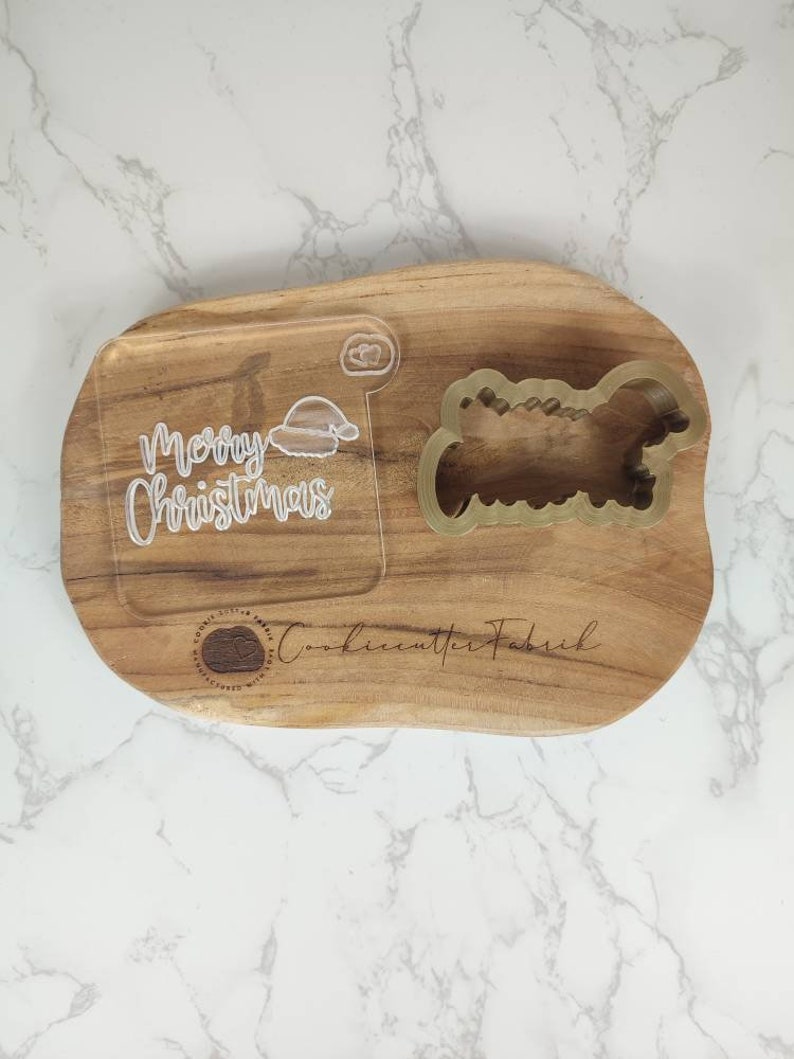 May include: A wooden cutting board with a "Merry Christmas" inscription and a Santa hat design. The board also features a cookie cutter in the shape of a Christmas tree. The text "Cookiecutter Fabrik" is engraved on the board.