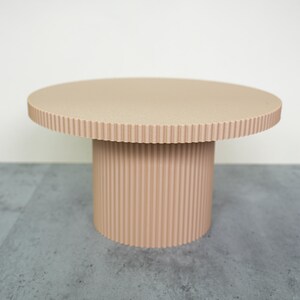 May include: A beige, round pedestal table with a textured surface and a ribbed cylindrical base. The table has a diameter of approximately 24 inches and stands about 18 inches tall. The design is modern and minimalist.