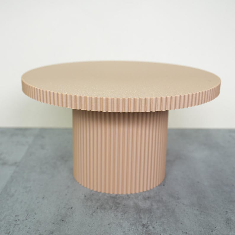 May include: A beige, round pedestal table with a textured surface and a ribbed cylindrical base. The table has a diameter of approximately 24 inches and stands about 18 inches tall. The design is modern and minimalist.