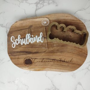 May include: A clear acrylic stamp featuring the white text Schulkind in a cursive font, paired with a gold-colored plastic cookie cutter shaped like a scalloped bone. The items are displayed on a natural wood board with a brand logo.