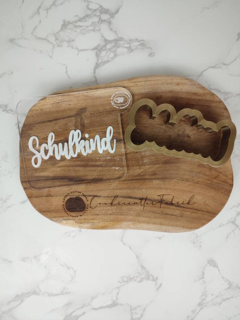 May include: A clear acrylic stamp featuring the white text Schulkind in a cursive font, paired with a gold-colored plastic cookie cutter shaped like a scalloped bone. The items are displayed on a natural wood board with a brand logo.