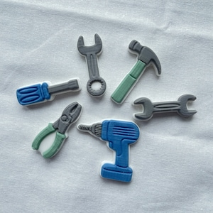 Könnte beinhalten: Ein Set aus sieben grauen und blauen Spielzeugwerkzeugen aus Kunststoff, darunter ein Schraubenschlüssel, ein Schraubendreher, ein Hammer, eine Zange, eine Bohrmaschine und zwei Schraubenschlüssel. Die Werkzeuge sind in einem Kreis auf einer weißen Oberfläche angeordnet.
