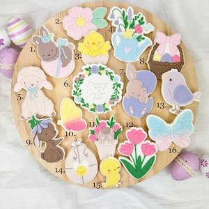 May include: A wooden platter displays Easter-themed decorations. These include bunny, chick, and lamb shapes, along with flowers, butterflies, and eggs. The words "Bunny Kisses Easter Wishes" are visible on one decoration. The decorations are in pastel colors.