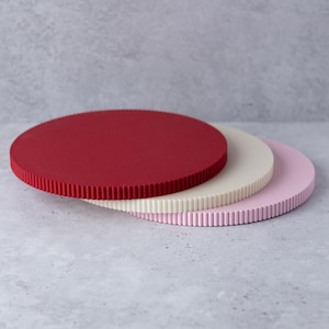 Op de afbeelding: Drie ronde taartborden in rood, crème en roze. Elk bord heeft een getextureerde, geribbelde rand. De borden zijn lichtjes gestapeld, waardoor hun verschillende kleuren en texturen worden getoond. De achtergrond is neutraal grijs.