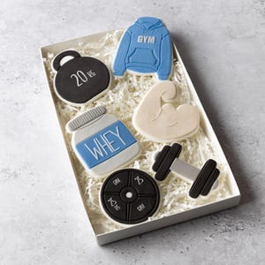 May include: A gift box containing gym-themed cookies. The cookies are shaped as a 20 kg weight, a blue hoodie with "GYM", a protein powder container with "WHEY", a flexing arm, a dumbbell, and a weight plate.