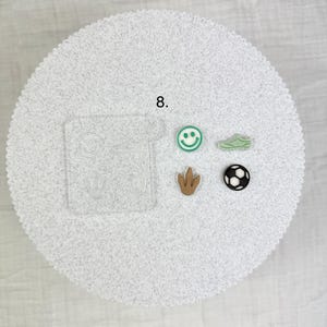 May include: A collection of small, colorful charms and a clear mold. The charms include a green smiley face, a green sneaker, a brown dinosaur footprint, and a black and white soccer ball. The number 8 is visible.
