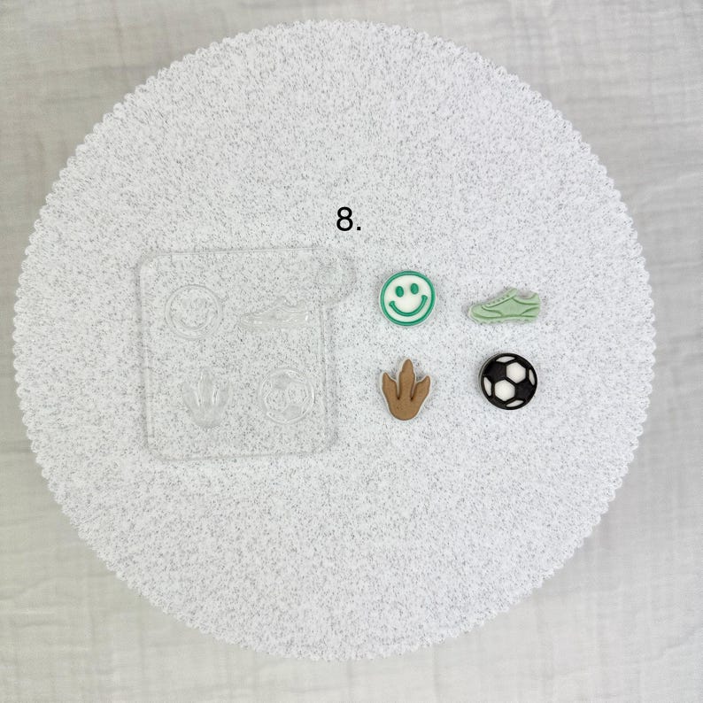 May include: A collection of small, colorful charms and a clear mold. The charms include a green smiley face, a green sneaker, a brown dinosaur footprint, and a black and white soccer ball. The number 8 is visible.