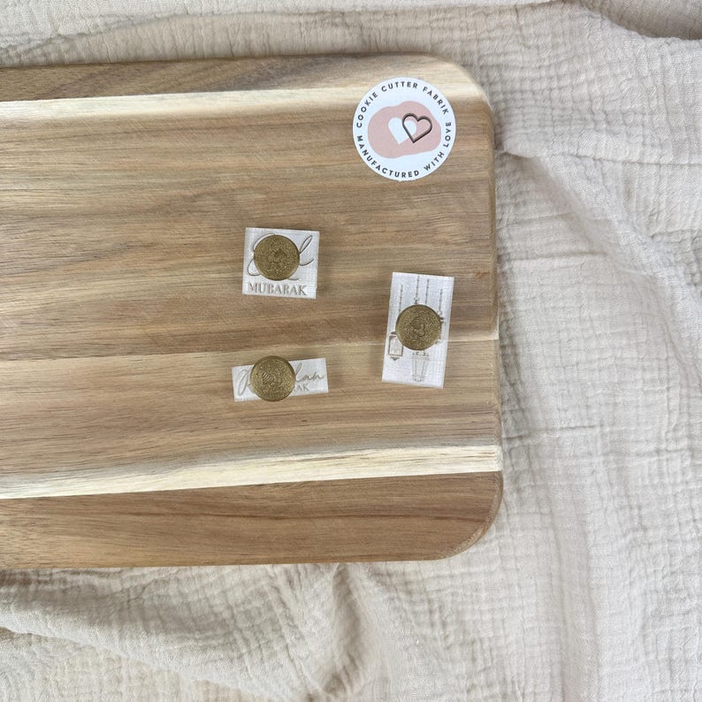 May include: Three cookie cutters on a wooden surface. The cutters are made of metal and have a gold finish. The cutters are shaped like a crescent moon, a star, and a lantern. The text "Mubarak" is printed on the crescent moon cutter.
