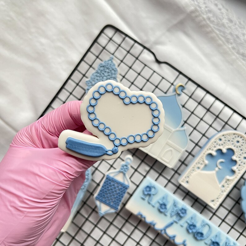 May include: A set of blue and white cookie cutters shaped like a mosque, a crescent moon, a lantern, and a string of prayer beads. The cutters are on a wire rack.