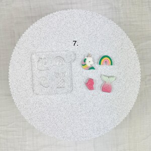 May include: A clear silicone mold with various shapes, including a pineapple, rainbow, and butterfly, alongside several colorful resin charms. The charms feature a unicorn, rainbow, butterfly, and mermaid tail. The number 7 is printed on the surface.