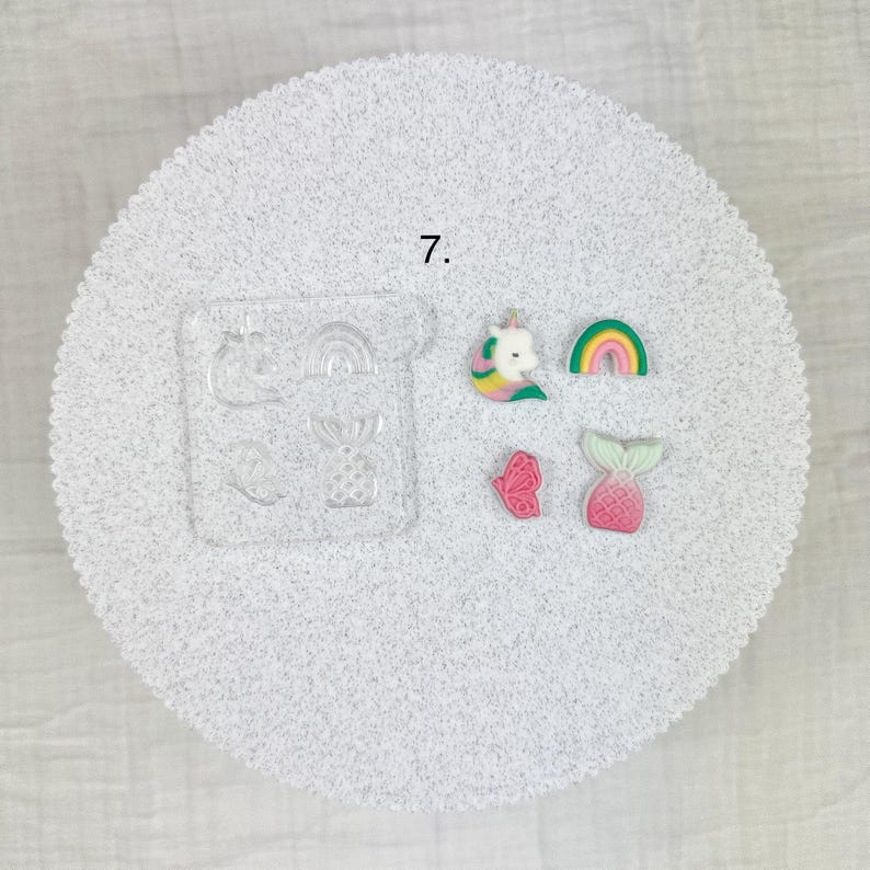 May include: A clear silicone mold with various shapes, including a pineapple, rainbow, and butterfly, alongside several colorful resin charms. The charms feature a unicorn, rainbow, butterfly, and mermaid tail. The number 7 is printed on the surface.