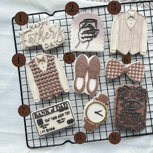 Op de afbeelding: Een verzameling Vaderdagkoekjes. De koekjes bevatten een "Happy Father's Day"-bericht, een hand die een drankje vasthoudt, een vest, een vlinderdas, schoenen, een creditcard, een horloge en een "Best Father"-plaquette.