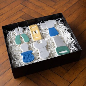 Könnte beinhalten: Eine schwarze Geschenkbox gefüllt mit weißen Papierschnipseln, die sechs Werkzeug-förmige Kekse enthält. Die Kekse sind in verschiedenen Farben bemalt, darunter Grau, Blau, Gelb und Grün. Die Werkzeuge umfassen eine Zange, ein Lineal, einen Hammer, einen Schraubenschlüssel und eine Säge.