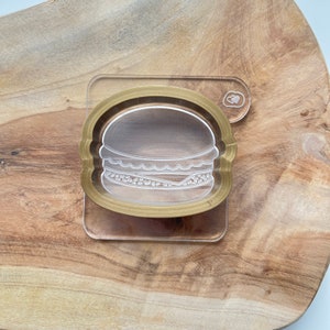 May include: A clear acrylic cookie cutter in the shape of a hamburger. The cutter is on a wooden surface.