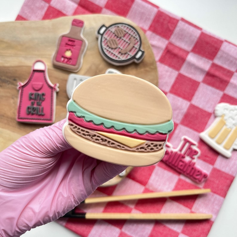 May include: A set of six colourful, decorated sugar cookies with a barbecue theme. The cookies include a hamburger, a grill, a beer mug, a bottle of hot sauce, a king of the grill apron, and a cookie with the text "The Grillfather".