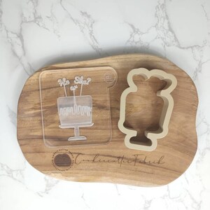 May include: A clear plastic cookie cutter with the text "He or She?" and a cake design. A beige silicone cookie cutter shaped like a cake is also shown on a wooden board.