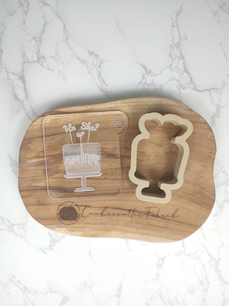 May include: A clear plastic cookie cutter with the text "He or She?" and a cake design. A beige silicone cookie cutter shaped like a cake is also shown on a wooden board.
