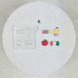 May include: A collection of school-themed embellishments, including the letters "ABC", numbers "123", a red apple, and a yellow pencil. A clear acrylic stamp with the words "ABC", "123", and an apple is also included. All items are on a white, textured surface.