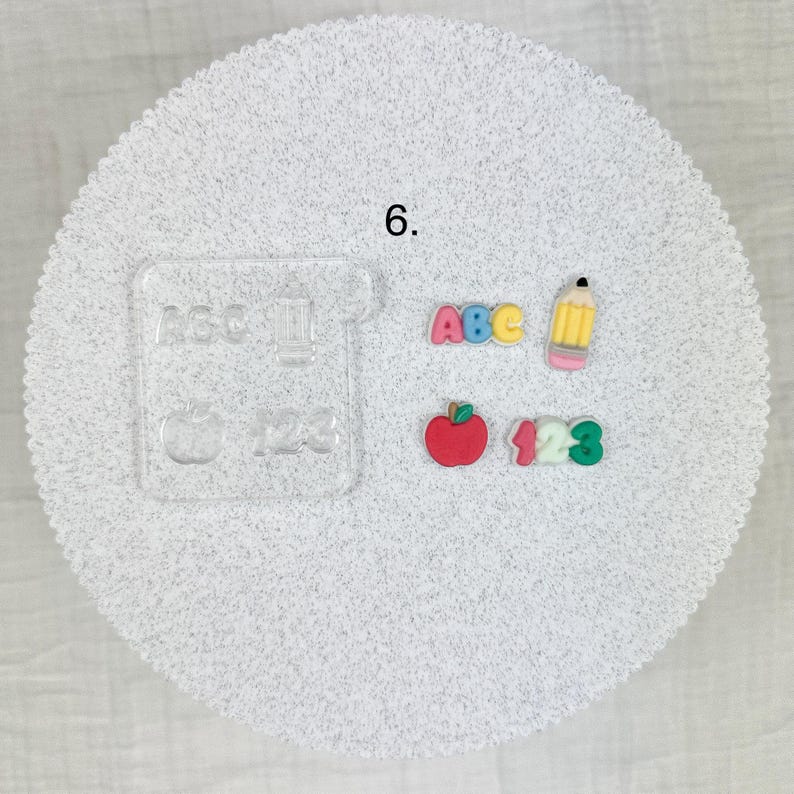 May include: A collection of school-themed embellishments, including the letters "ABC", numbers "123", a red apple, and a yellow pencil. A clear acrylic stamp with the words "ABC", "123", and an apple is also included. All items are on a white, textured surface.