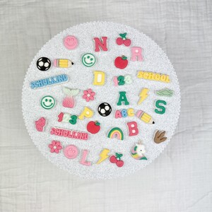 May include: A collection of colorful, decorative embellishments, including letters, numbers, and various shapes like a soccer ball, pencil, and smiley faces. The word "SCHOOLKIND" is visible. The items are arranged on a white, textured surface.