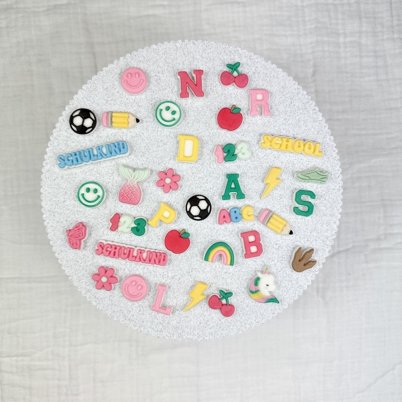 May include: A collection of colorful, decorative embellishments, including letters, numbers, and various shapes like a soccer ball, pencil, and smiley faces. The word "SCHOOLKIND" is visible. The items are arranged on a white, textured surface.