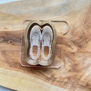 May include: A clear acrylic coaster with a gold outline and a pair of sneakers etched inside. The coaster has a small heart shape in the top right corner.