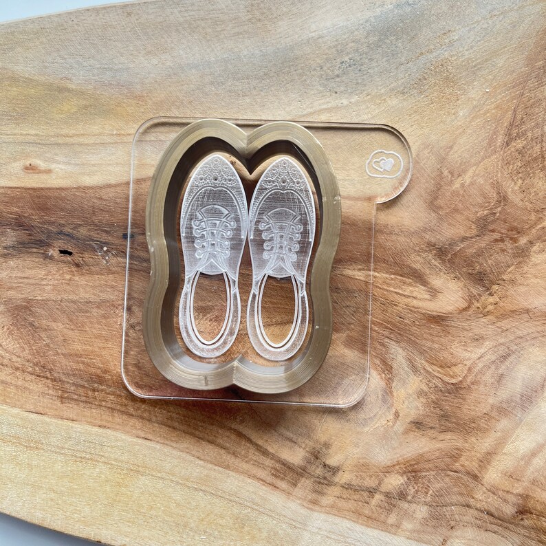 May include: A clear acrylic coaster with a gold outline and a pair of sneakers etched inside. The coaster has a small heart shape in the top right corner.