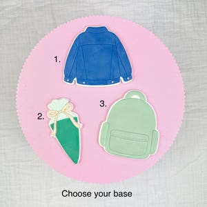 May include: A selection of three die-cut shapes on a pink background: a blue jacket, a green gift bag, and a green backpack. The text "Choose your base" is at the bottom. These shapes are likely for crafting or design projects.