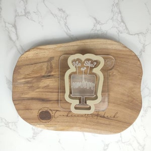 May include: A clear plastic cookie cutter shaped like a cake with the words "He or She?" on top. The cutter is on a wooden surface.