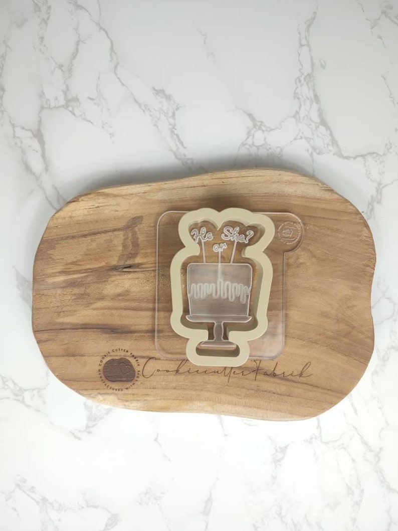 May include: A clear plastic cookie cutter shaped like a cake with the words "He or She?" on top. The cutter is on a wooden surface.
