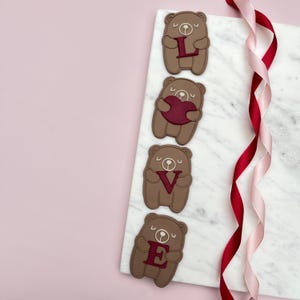 Puede incluir: Cuatro decoraciones en forma de oso marrón que deletrean "LOVE" con letras rojas y un corazón rojo. Los osos están dispuestos sobre una superficie de mármol blanco con cintas rosas y rojas. El fondo es rosa claro.