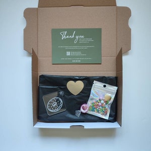 May include: An open cardboard box with a black tissue paper lining. The box contains a clear acrylic donut charm, a gold heart charm, a small bag of colorful sprinkles, and a thank you card.