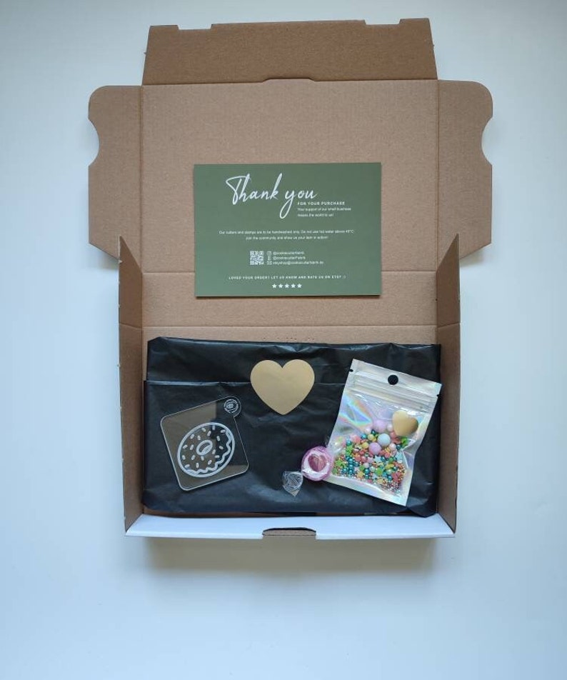 May include: An open cardboard box with a black tissue paper lining. The box contains a clear acrylic donut charm, a gold heart charm, a small bag of colorful sprinkles, and a thank you card.